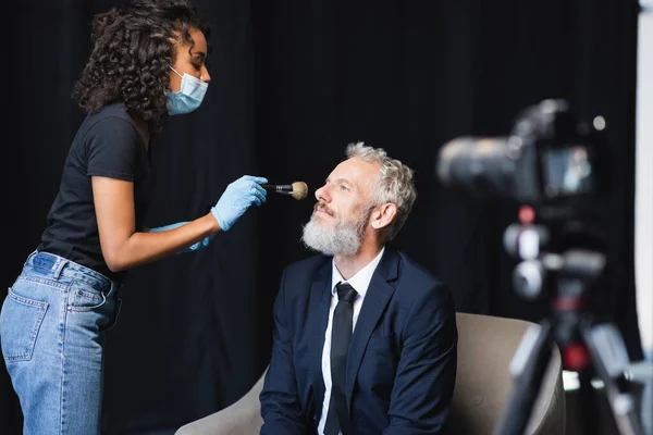 Artista de maquillaje afroamericano en máscara médica y guantes de látex aplicando polvo facial en hombre de negocios cerca de la cámara digital borrosa - foto de stock