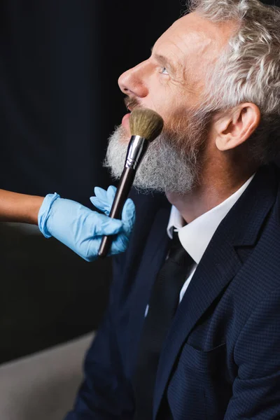 Maquilleur afro-américain en gant de latex appliquant de la poudre visage sur un homme d'affaires en costume — Photo de stock
