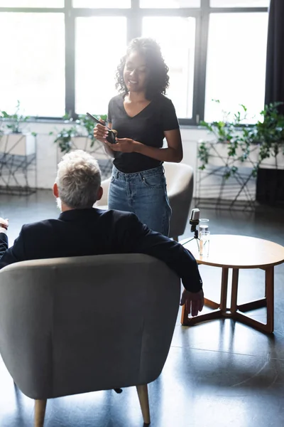 Joyeux maquilleur afro-américain debout près de l'homme d'affaires en studio d'interview — Photo de stock