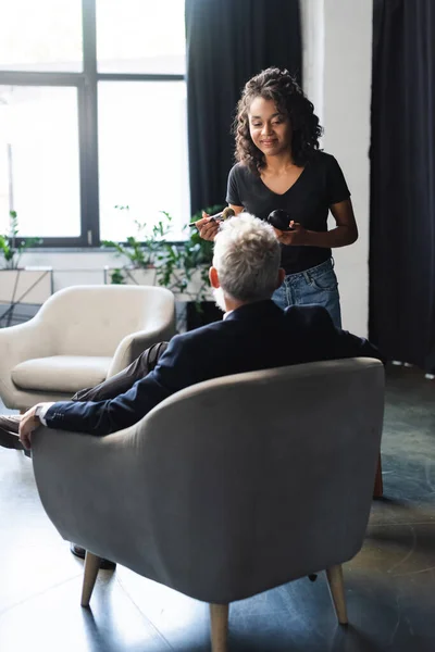 Sonriente afroamericano maquillaje artista de pie cerca de hombre de negocios en estudio de entrevista - foto de stock