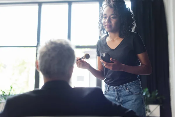 Maquilleur afro-américain tenant un pinceau cosmétique près d'un homme d'affaires flou dans un studio d'interview — Photo de stock