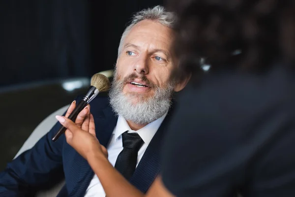 Maquilleur afro-américain flou appliquant de la poudre sur le visage de l'homme d'affaires en studio d'interview — Photo de stock