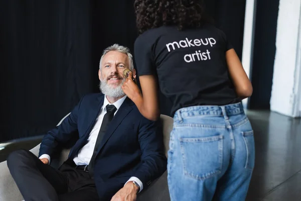 Encaracolado artista de maquiagem afro-americano em t-shirt com letras aplicando pó facial em empresário feliz em estúdio de entrevista — Fotografia de Stock