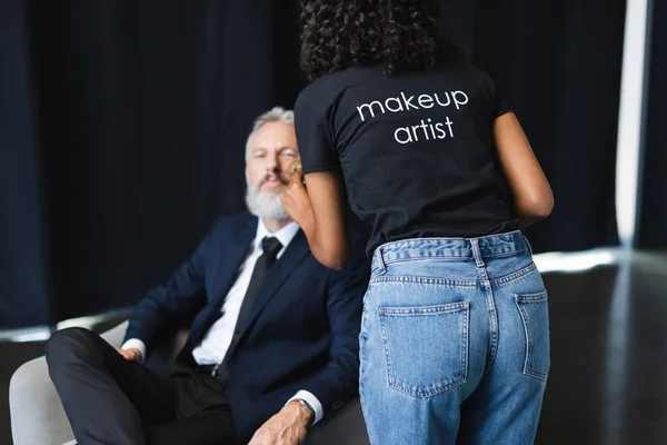 African american makeup artist in t-shirt with lettering applying face powder on blurred businessman in interview studio — Stock Photo