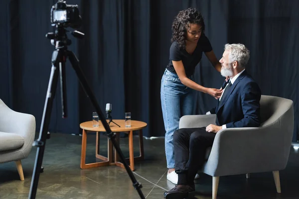 Morena afro-americana assistente ajustando microfone em terno de empresário em estúdio de entrevista — Fotografia de Stock