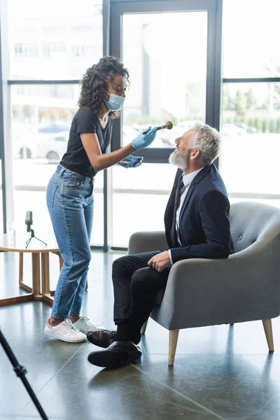 African american makeup artist in medical mask and latex gloves applying face powder on businessman in interview studio — Stock Photo