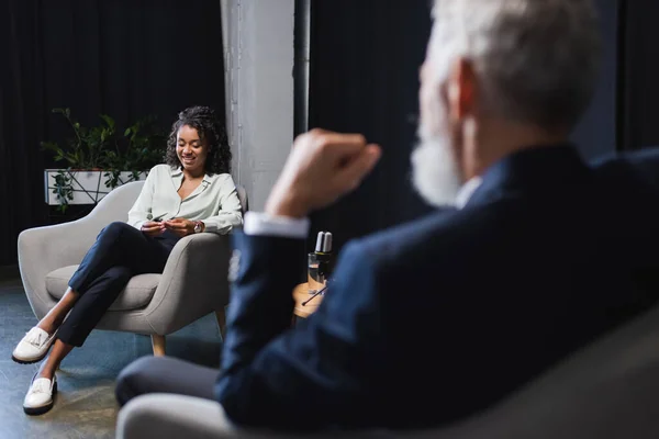 Happy african american journalist smiling near blurred businessman during talk show — Stock Photo