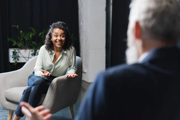 Cheerful african american journalist gesturing and looking at blurred businessman during talk show — Stock Photo