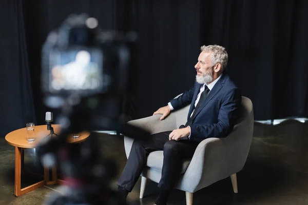 Homme d'affaires en costume assis dans un fauteuil lors d'une interview près d'un appareil photo numérique flou — Photo de stock