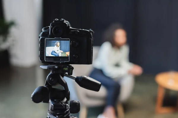 Lockige afrikanisch-amerikanische Journalistin sitzt im Sessel neben verschwommener Digitalkamera — Stockfoto