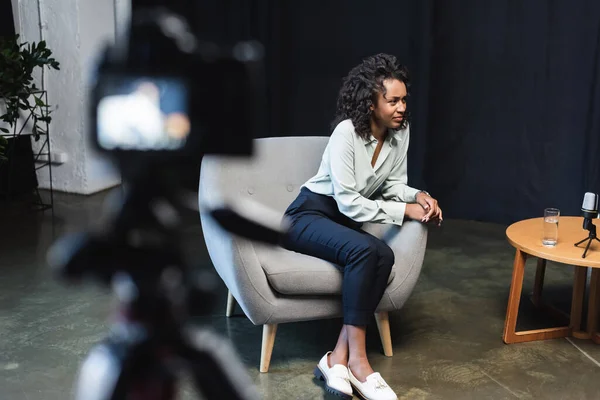 Journaliste afro-américain bouclé assis dans un fauteuil près de la table basse et appareil photo numérique flou — Photo de stock