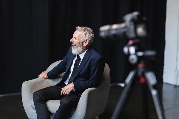 Alegre hombre de negocios en traje riendo mientras se sienta en sillón durante la entrevista cerca de la cámara digital borrosa en el trípode - foto de stock