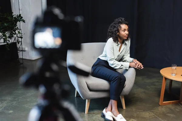 Morena africano americano periodista sentado en sillón cerca de mesa de café y borrosa cámara digital - foto de stock