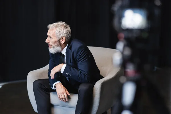 Homme d'affaires en costume assis dans un fauteuil gris lors d'une interview près d'un appareil photo numérique flou en studio — Photo de stock