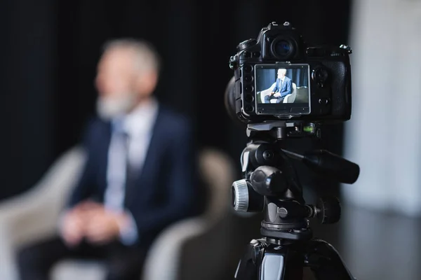 Digital camera with businessman in suit sitting in grey armchair during interview on screen — Stock Photo