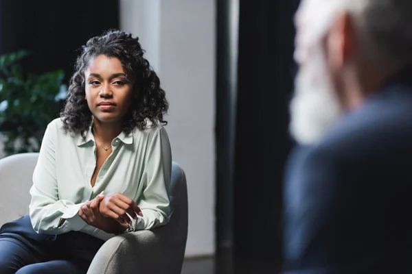 Brunette african american journalist sitting in armchair and looking at blurred guest — Stock Photo