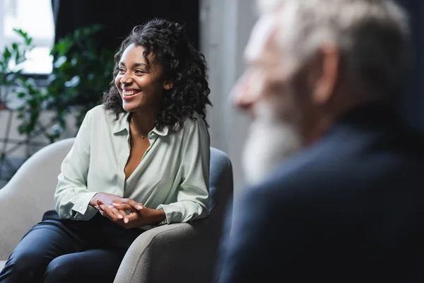 Cheerful african american journalist sitting in armchair near blurred guest — Stock Photo