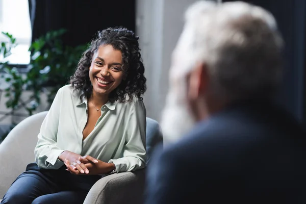 Journaliste afro-américain positif assis dans un fauteuil et souriant près invité flou — Photo de stock