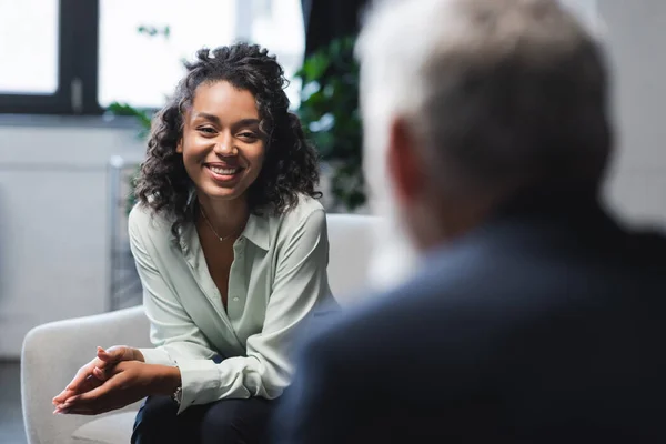 Joyful african american journalist sitting in armchair and smiling near blurred guest — Stock Photo