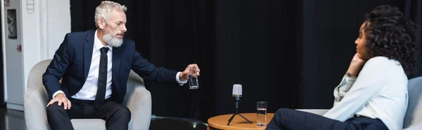 Homme d'affaires en costume prendre un verre d'eau lors d'une interview près d'un journaliste afro-américain, bannière — Photo de stock