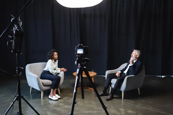 Periodista afroamericano rizado sentado en sillón y hablando con el invitado en el estudio - foto de stock