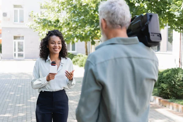 Cámara borrosa con cámara de vídeo cerca de reportero afroamericano feliz con micrófono haciendo reportaje - foto de stock