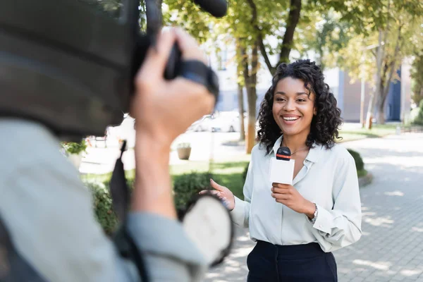 Розмитий оператор з відеокамерою біля усміхненого афроамериканського репортера з мікрофоном роблять репортаж — стокове фото