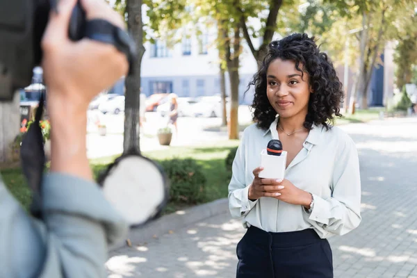 Cámara borrosa con cámara de vídeo cerca de reportero afroamericano bastante con micrófono haciendo reportaje - foto de stock