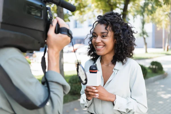 Verschwommener Kameramann mit Videokamera in der Nähe lächelnder afrikanisch-amerikanischer Journalist mit Mikrofon für Reportagen — Stockfoto