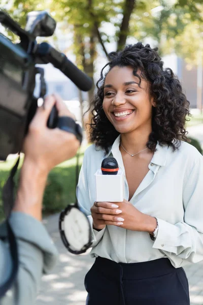 Sonriente periodista afroamericano con micrófono haciendo reportaje cerca de cámara borrosa con cámara de video - foto de stock