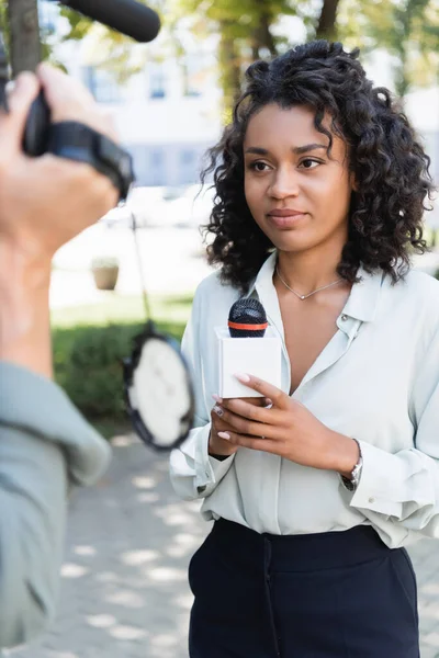 Bastante afroamericano periodista con micrófono haciendo reportaje cerca de cámara borrosa con cámara de vídeo - foto de stock