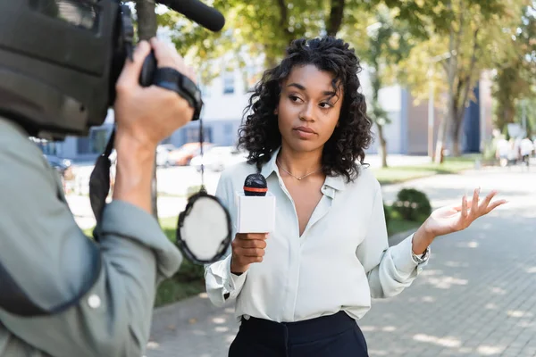 Curly african american journalist with microphone pointing with hand while doing reportage near blurred cameraman — Stock Photo