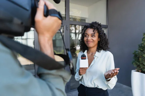 Щасливий афроамериканський журналіст з жестом мікрофона під час репортажу біля розмитого оператора — стокове фото