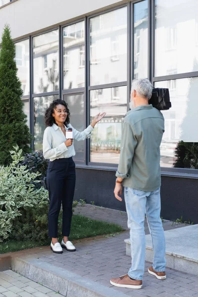 Full length of cheerful african american journalist pointing with hand at building while doing reportage near cameraman — Stock Photo