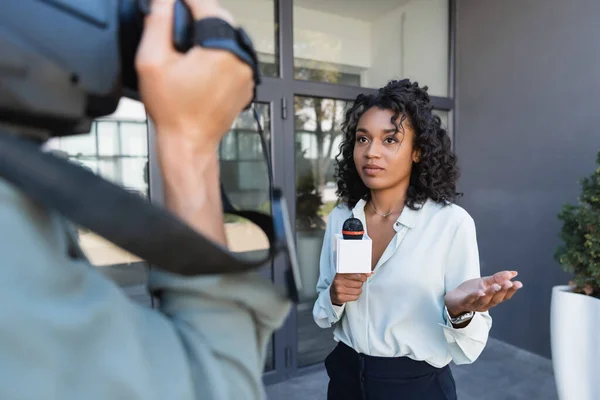 Periodista afroamericano bastante con el gesto del micrófono mientras que hace el reportaje cerca del camarógrafo borroso - foto de stock