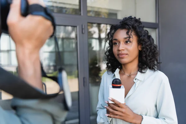 Curly african american journalist with microphone gesturing while doing reportage near blurred cameraman — Stock Photo