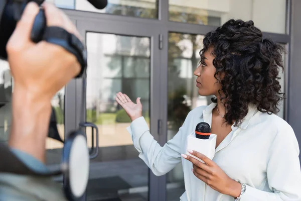Periodista afroamericano señalando con la mano en el edificio mientras hace reportaje cerca de cámara borrosa - foto de stock