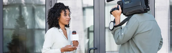 Morena afroamericana periodista haciendo reportaje cerca de camarógrafo fuera, pancarta - foto de stock