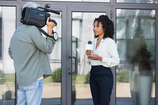 Brünette afrikanisch-amerikanische Journalistin macht Reportagen in der Nähe des Operators mit Videokamera draußen — Stockfoto