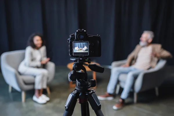 Cámara digital con hombre de negocios en traje sentado en sillón gris durante entrevista con periodista afroamericano en pantalla - foto de stock