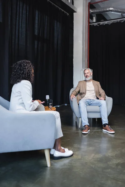 Ricci afroamericani giornalista in giacca e cravatta parlare con uomo d'affari barbuto seduto in poltrona durante l'intervista — Foto stock