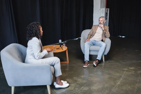 Lockige afrikanisch-amerikanische Journalistin im Anzug im Gespräch mit einem Geschäftsmann im Sessel während eines Interviews — Stockfoto