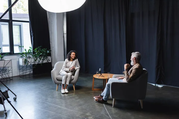 Positivo giornalista afroamericano in giacca e cravatta parlare con uomo d'affari seduto in poltrona durante l'intervista — Foto stock