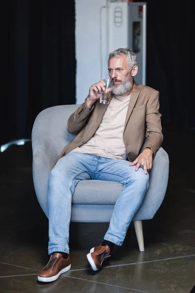 Homem de negócios barbudo segurando vidro de água e sentado em poltrona cinza durante a entrevista — Fotografia de Stock