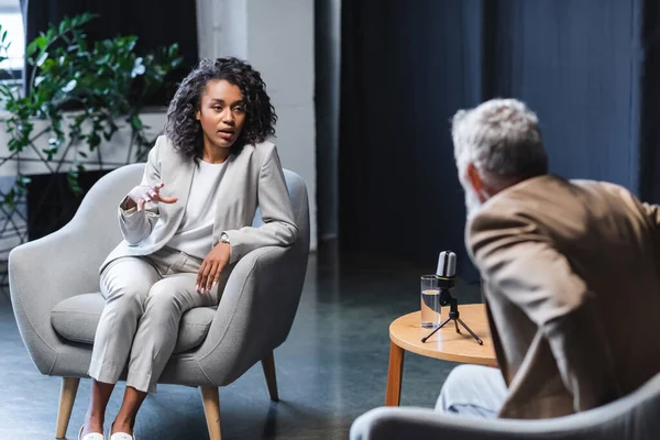 African american journalist gesturing while talking with blurred businessman sitting in armchair during interview — Stock Photo