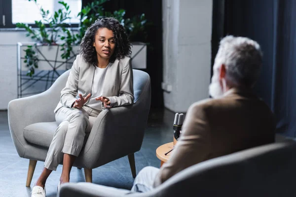 Encaracolado jornalista afro-americano gesticulando enquanto conversava com empresário desfocado sentado em poltrona durante a entrevista — Fotografia de Stock