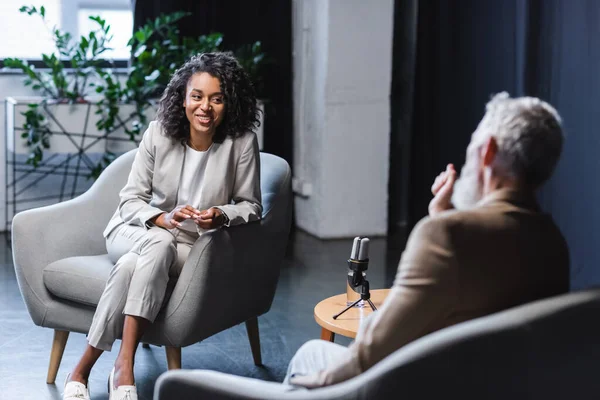 Feliz periodista afroamericano haciendo un gesto mientras habla con un hombre de negocios borroso sentado en un sillón durante la entrevista - foto de stock
