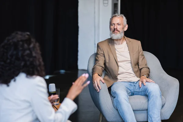 Flou journaliste afro-américain gestuelle tout en parlant avec un homme d'affaires assis dans le fauteuil pendant l'interview — Photo de stock