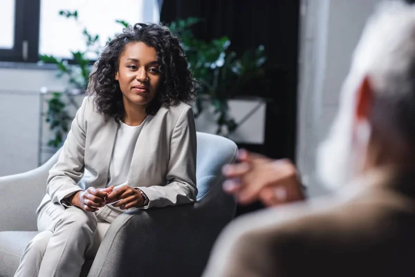 Ziemlich afrikanisch-amerikanischer Journalist blickt auf verschwommenen Geschäftsmann, der im Sessel sitzt und gestikuliert, während er während des Interviews spricht — Stockfoto