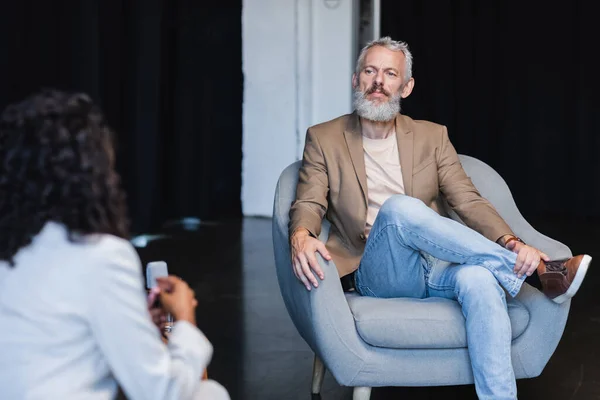 Jornalista afro-americano desfocado conversando com empresário confiante sentado em poltrona durante a entrevista — Fotografia de Stock